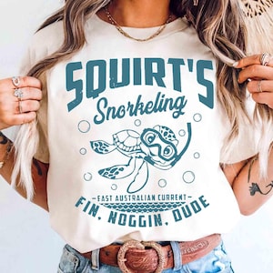 May include: A cream-colored t-shirt featuring a blue graphic of a sea turtle wearing a snorkel mask. The text reads SQUIRT'S Snorkeling, FAST AUSTRALIAN CURRENT, and FIN, NOGGIN, DUDE, surrounded by small blue bubbles on the front of the garment.
