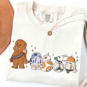 May include: Cream-colored t-shirt featuring a cartoon illustration of Chewbacca and several droids from Star Wars. The design includes R2-D2, BB-8, and other droid characters. The shirt has a crew neck and short sleeves.