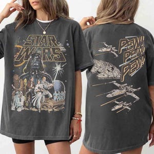 Pode incluir: Uma t-shirt de manga curta cinzento-carvão com um gráfico de Star Wars na frente com Darth Vader, Luke Skywalker, Princesa Leia, Chewbacca e droides. As costas exibem caças X-wing e o texto Pew! Pew! Pew! em dourado.