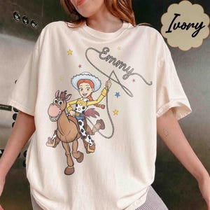 May include: A cream-colored short-sleeve t-shirt featuring a graphic of a cowgirl riding a brown horse. The cowgirl wears a white hat, yellow shirt, and blue pants. A lasso swirls above with the name Emmy written in cursive script and small stars.
