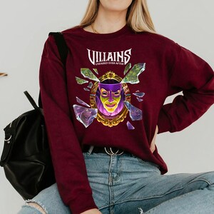 Puede incluir: Sudadera burdeos de manga larga con el texto "Villains Unfairly Ever After". El gr&aacute;fico presenta un espejo dorado con una m&aacute;scara morada y amarilla, rodeado de cristales rotos. Se ve una mochila negra.