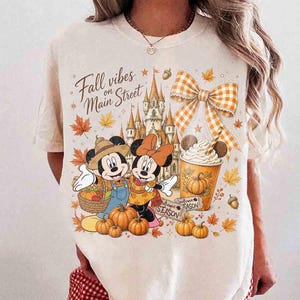 May include: A cream-colored t-shirt featuring a graphic print with the text Fall vibes on Main Street. The design includes cartoon mice, a castle, a large orange and white checkered bow, pumpkins, autumn leaves, and a cup with whipped cream.