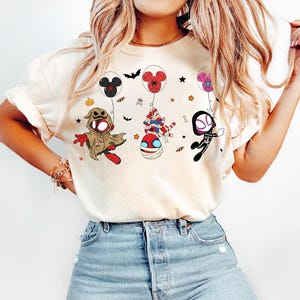 Puede incluir: Camiseta color crema con personajes de dibujos animados de Halloween, incluyendo a Oogie Boogie, Spider-Man momificado y Spider-Gwen, cada uno sostenido por globos con forma de Mickey Mouse. El dise&ntilde;o incluye murci&eacute;lagos, estrellas y una calabaza.