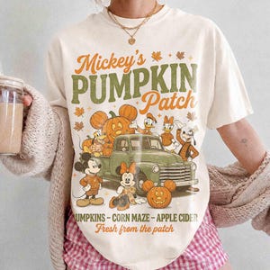 Könnte beinhalten: Ein cremefarbenes T-Shirt mit einem Grafikdruck und dem Text Mickey's PUMPKIN Patch. Das Design zeigt einen alten grünen Pickup, verschiedene Zeichentrickfiguren, Kürbisse und Herbstblätter sowie den Text PUMPKINS - CORN MAZE - APPLE CIDER Fresh from the patch.
