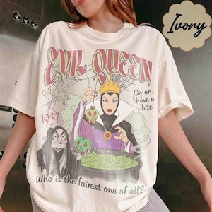 May include: A cream-coloured t-shirt featuring a graphic print of a queen in a purple robe and a crown, holding a green apple over a cauldron. The text reads Evil Queen, 1937, Go on have a bite, and Who is the fairest one of all? A small witch stands nearby.