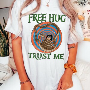 May include: White t-shirt with a graphic featuring a cartoon character wrapped by a snake, set against a blue and orange spiral background. The text "FREE HUG" and "TRUST ME" are printed in green.