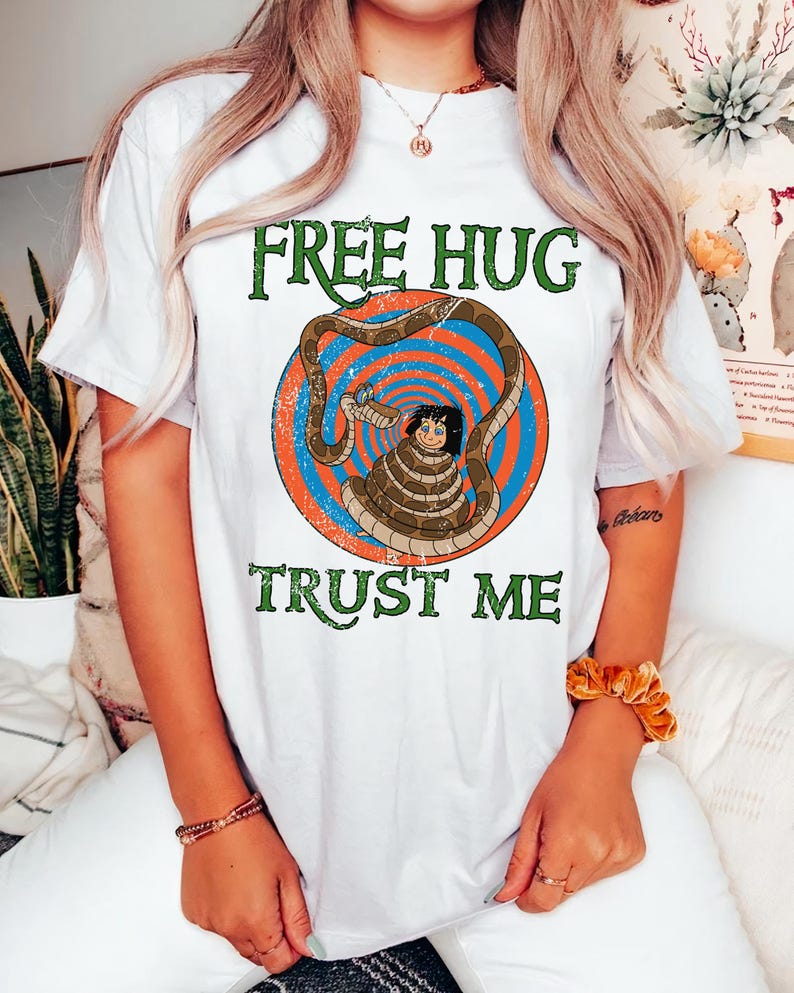 May include: White t-shirt with a graphic featuring a cartoon character wrapped by a snake, set against a blue and orange spiral background. The text "FREE HUG" and "TRUST ME" are printed in green.