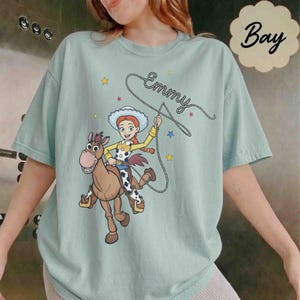 May include: A light green t-shirt featuring a graphic of the character Jessie from Toy Story riding Bullseye the horse. The design includes a lasso, small stars, and the name Emmy written in cursive script. The shirt has a crew neckline.