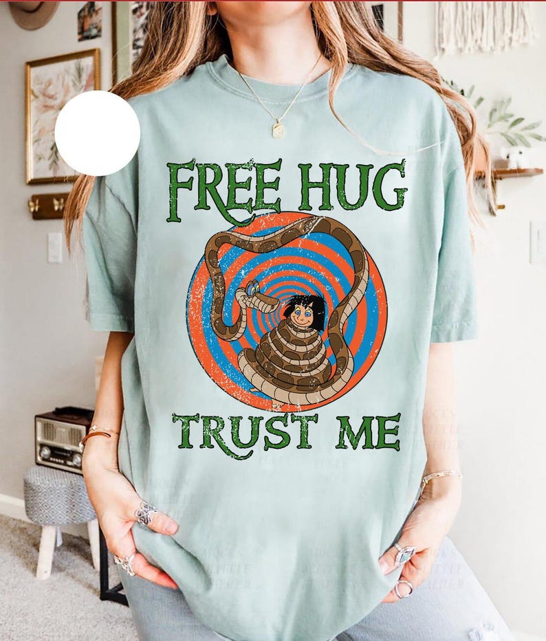 May include: A light blue t-shirt with the text "FREE HUG TRUST ME" in green. The shirt features a graphic of a person wrapped in a snake, inside a red and blue circle. The shirt has short sleeves.