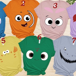 May include: A collection of seven colorful graphic t-shirts featuring cartoon character faces. The shirts are in various colors, including blue, orange, pink, purple, yellow, green, and gray. Each shirt has a unique design, perfect for fans of animated movies.