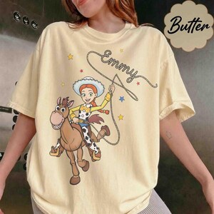 May include: A pale yellow t-shirt featuring a graphic of a cowgirl character with red hair and a white hat riding a brown horse. The character holds a lasso that forms the name Emmy in cursive script. Small yellow and blue stars surround the design.