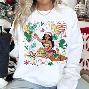 K&ouml;nnte beinhalten: Wei&szlig;es Sweatshirt mit einer farbenfrohen Grafik eines Cartoon-Charakters mit Weihnachtsm&uuml;tze, umgeben von Luftballons, Geschenken und festlichen Dekorationen. Das Design umfasst ein Schwein, ein Huhn und Mickey Mouse-f&ouml;rmige Luftballons. Ein grafisches T-Shirt zum Thema Feiertage.