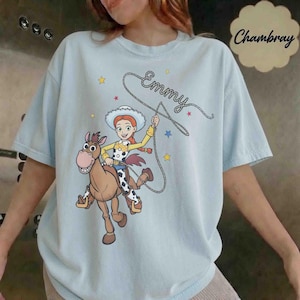 May include: A light blue t-shirt featuring a graphic of a cowgirl character riding a brown horse. The cowgirl holds a lasso with the name Emmy written in cursive script above her. Small yellow and blue stars are scattered around the design.