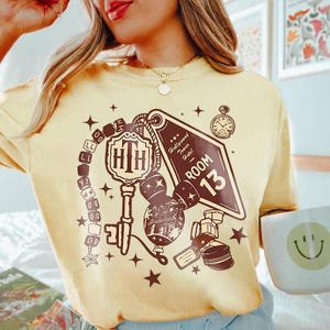 Könnte beinhalten: Hellgelbes T-Shirt mit braunem Grafikdesign. Das Design umfasst einen Schlüssel, einen Hotelzimmer-Schlüsselanhänger mit der Aufschrift "Hollywood Tower Hotel Room 13" und andere thematische Elemente. Das Shirt hat auch eine Taschenuhr und Sterne.
