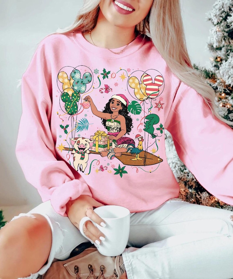 K&ouml;nnte beinhalten: Rosafarbenes Sweatshirt mit einer Cartoon-Illustration einer Frau mit Weihnachtsm&uuml;tze, umgeben von festlichen Elementen. Mickey Mouse-f&ouml;rmige Luftballons, ein Schwein und ein Hahn sind ebenfalls abgebildet. Das Design hat ein Feiertags-Thema.