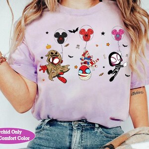 Puede incluir: Una camiseta de color lila claro con personajes de dibujos animados disfrazados para Halloween, flotando con globos con forma de Mickey Mouse. La camiseta incluye el texto "Orchid Only With Comfort Color". Murci&eacute;lagos, estrellas y calabazas est&aacute;n dispersos.