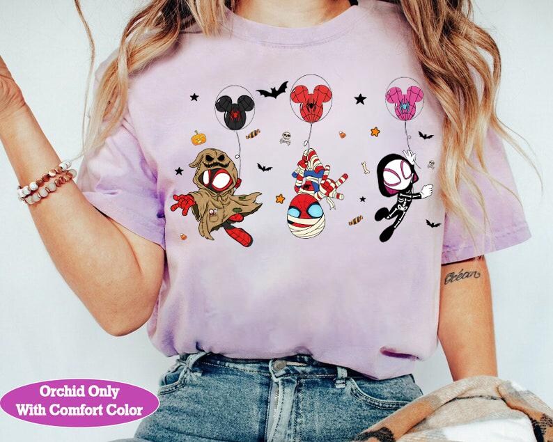 Puede incluir: Una camiseta de color lila claro con personajes de dibujos animados disfrazados para Halloween, flotando con globos con forma de Mickey Mouse. La camiseta incluye el texto "Orchid Only With Comfort Color". Murci&eacute;lagos, estrellas y calabazas est&aacute;n dispersos.