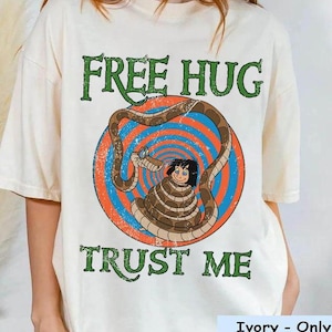May include: Ivory-colored t-shirt with a graphic featuring a cartoon snake coiled around a person. The text "FREE HUG" and "TRUST ME" are printed in green. The graphic is set against a blue and orange circular background.