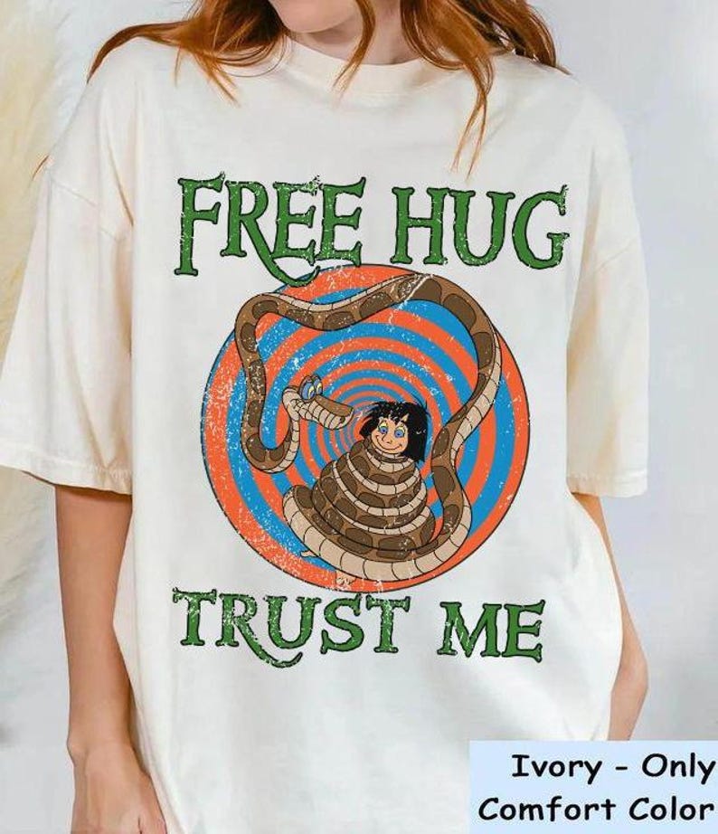 May include: Ivory-colored t-shirt with a graphic featuring a cartoon snake coiled around a person. The text "FREE HUG" and "TRUST ME" are printed in green. The graphic is set against a blue and orange circular background.