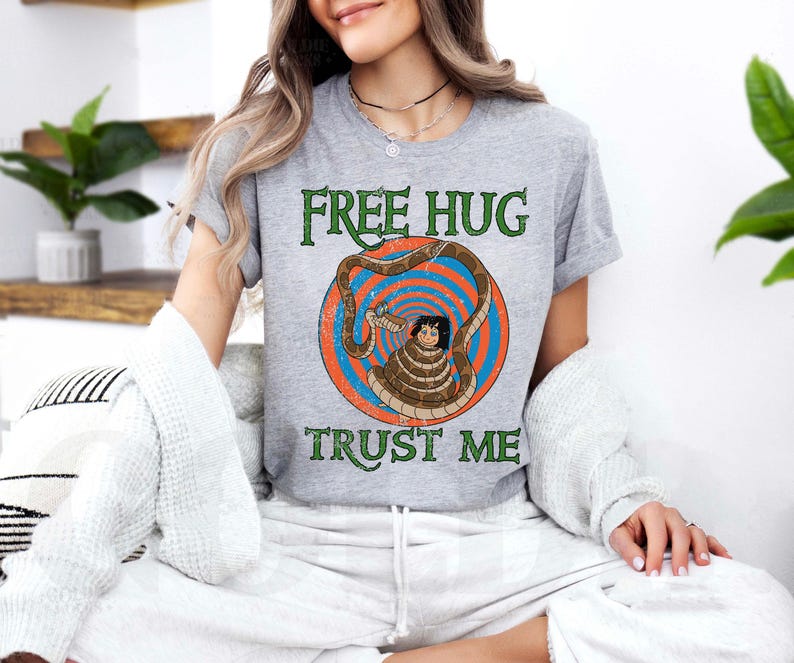 May include: A gray t-shirt with the words "FREE HUG TRUST ME" in green text. The shirt features a cartoon illustration of a person wrapped in a snake. The illustration is set within a blue and orange circular design.