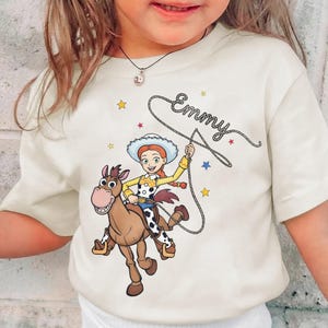 May include: A cream-colored t-shirt featuring a graphic of a cowgirl with red hair and a white hat riding a brown horse. The cowgirl holds a lasso that forms the name Emmy in cursive script. Small colorful stars are scattered around the design.
