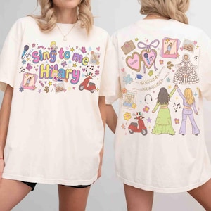 May include: An off-white t-shirt featuring a graphic print on the front and back. The front displays the text "Sing to me Hilary" with colorful illustrations of a scooter, stars, and hearts. The back features illustrations of two figures, a scooter, and the text "Lizzie McGuire".
