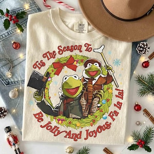 May include: A cream-colored t-shirt featuring a graphic of two green frogs inside a Christmas wreath. One frog wears a top hat and the other holds a crutch. The text reads, Tis The Season To Be Jolly And Joyous. Fa la la! The shirt has a Comfort Colors label.