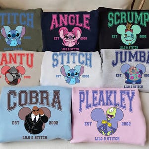May include: Nine different coloured sweatshirts featuring a variety of cartoon characters from the Disney film Lilo & Stitch. Each sweatshirt features a different character, including Stitch, Angle, Scrump, Gantu, Jumba, Cobra, and Pleakley. The sweatshirts are all in a college-style design with the character's name and the year 2002 printed on them.