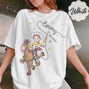 May include: A white t-shirt featuring a graphic of a cowgirl character riding a brown horse. The character holds a lasso that forms the name Emmy in cursive script. The design includes small yellow, red, and blue stars scattered around the figures.