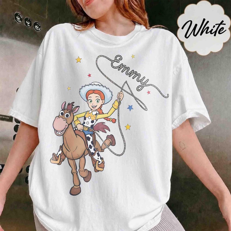 May include: A white t-shirt featuring a graphic of a cowgirl character riding a brown horse. The character holds a lasso that forms the name Emmy in cursive script. The design includes small yellow, red, and blue stars scattered around the figures.