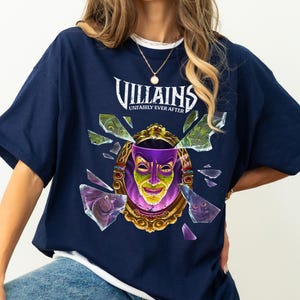 Puede incluir: Camiseta azul marino con un dise&ntilde;o de un espejo dorado con una cara morada y verde, rodeado de cristales rotos. El texto "Villains Unfairly Ever After" est&aacute; arqueado sobre el espejo.