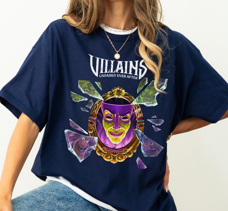 Puede incluir: Camiseta azul marino con un dise&ntilde;o de un espejo dorado con una cara morada y verde, rodeado de cristales rotos. El texto "Villains Unfairly Ever After" est&aacute; arqueado sobre el espejo.