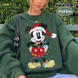 Puede incluir: Una sudadera verde bosque con un gráfico de Mickey Mouse usando un gorro de Santa rojo y blanco. Mickey está rodeado de luces navideñas de colores. La sudadera tiene mangas largas y cuello redondo. El texto en la imagen dice SWEATSHIRT Forest Green.