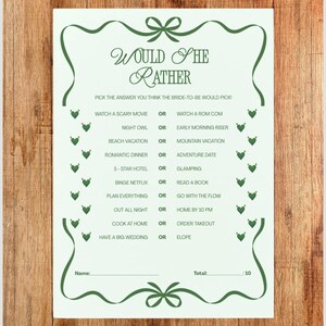 May include: A white paper game card with green decorative elements and text. The card reads "Would She Rather" and includes a series of questions for a bridal shower game. The card is framed with green bows and decorative elements.