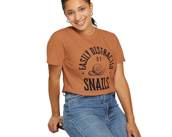 May include: A rust-colored t-shirt with the text "EASILY DISTRACTED BY SNAILS" in black. The design includes a snail illustration. The person in the image is wearing blue jeans.