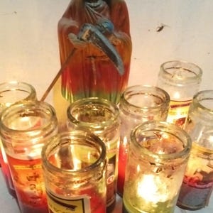 May include: A collection of seven cylindrical glass prayer candles with lit wicks, arranged in a cluster. Each candle contains colored wax and a label. Behind the candles stands a figurine of a hooded skeletal figure wearing a red robe.