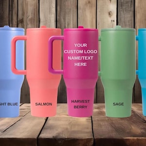 K&ouml;nnte beinhalten: F&uuml;nf hohe Isolierbecher mit Henkel in den Farben Hellblau, Lachs, Beere, Salbei und Gletscher. Der mittlere beerenfarbene Becher tr&auml;gt den Text YOUR CUSTOM LOGO NAME/TEXT HERE. Jeder Becher ist mit einem passenden Deckel und Henkel versehen.