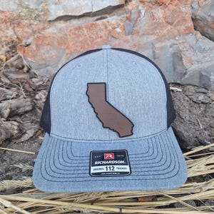 May include: A gray and black trucker hat with a brown leather California state outline patch. The hat has an adjustable strap and a black mesh back. The hat has the text "Richardson" and "Original Trucker" on the front.