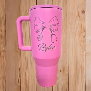 Puede incluir: Un termo de viaje rosa con asa y tapa. El termo presenta un dise&ntilde;o de lazo y estetoscopio, con el nombre "Rylee" en escritura cursiva. El termo es de acero inoxidable y est&aacute; dise&ntilde;ado para mantener las bebidas calientes o fr&iacute;as.