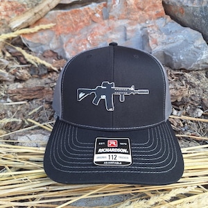 May include: Black and gray trucker hat with a black silhouette of a rifle on the front. The hat has a black and gray mesh back and a black adjustable strap with a white label that reads "EST. 1970 RICHARDSON. ORIGINAL 112 TRUCKER ADJUSTABLE".
