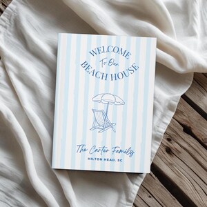 May include: A hardcover book with a light blue and white striped pattern. The cover reads "WELCOME To Our BEACH HOUSE" in a circular design, with an illustration of a beach umbrella and chair. Below, it says "The Carter Family, Hilton Head, SC."
