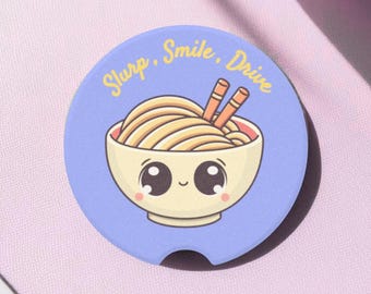 May include: A round, light blue car coaster featuring a cartoon bowl of ramen with smiling eyes and chopsticks. The text "Slurp, Smile, Drive" is written in yellow cursive around the edge of the coaster.