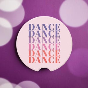 May include: A round, off-white car coaster with a curved notch. The word "DANCE" is printed vertically in a gradient of colors, transitioning from blue to pink to red. The coaster is set against a purple background with blurred white circles.