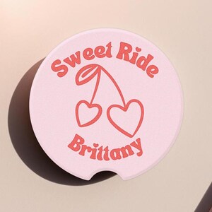 May include: A light pink car coaster with the words "Sweet Ride Brittany" in a retro font. A red cherry graphic with heart-shaped cherries is in the center. The coaster is round with a small notch.