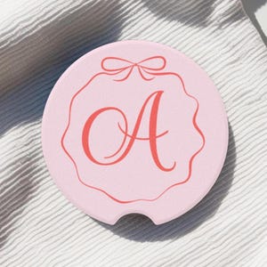 Op de afbeelding: Ronde roze onderzetter met een rode rand en een rode strik bovenaan. De letter "A" staat in het midden van de onderzetter in rood.