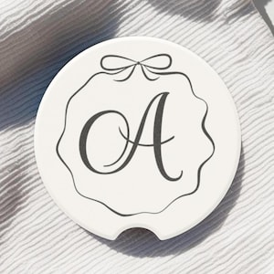 May include: White ceramic coaster with a black outline of a decorative frame with a bow at the top and the letter "A" in a script font inside the frame.