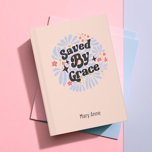 May include: A pink notebook with the text "Saved By Grace" in black lettering surrounded by blue and pink flowers. The name "Mary Anne" is printed at the bottom of the notebook.