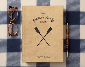 May include: A brown guest book with the text "The Jackson Family Cabin" and two crossed oars. The book is on a blue and white checked blanket.