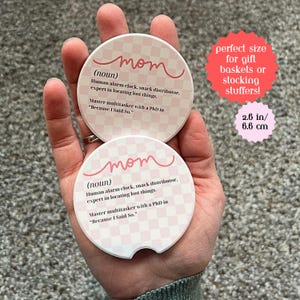 May include: Two round pink and white checkered coasters with the word "mom" written in pink cursive. The text below reads "(noun) Human alarm clock, snack distributor. expert in locating lost things. Master multitasker with a PhD in "Because I Said So."". The coasters are 2.6 inches in diameter.