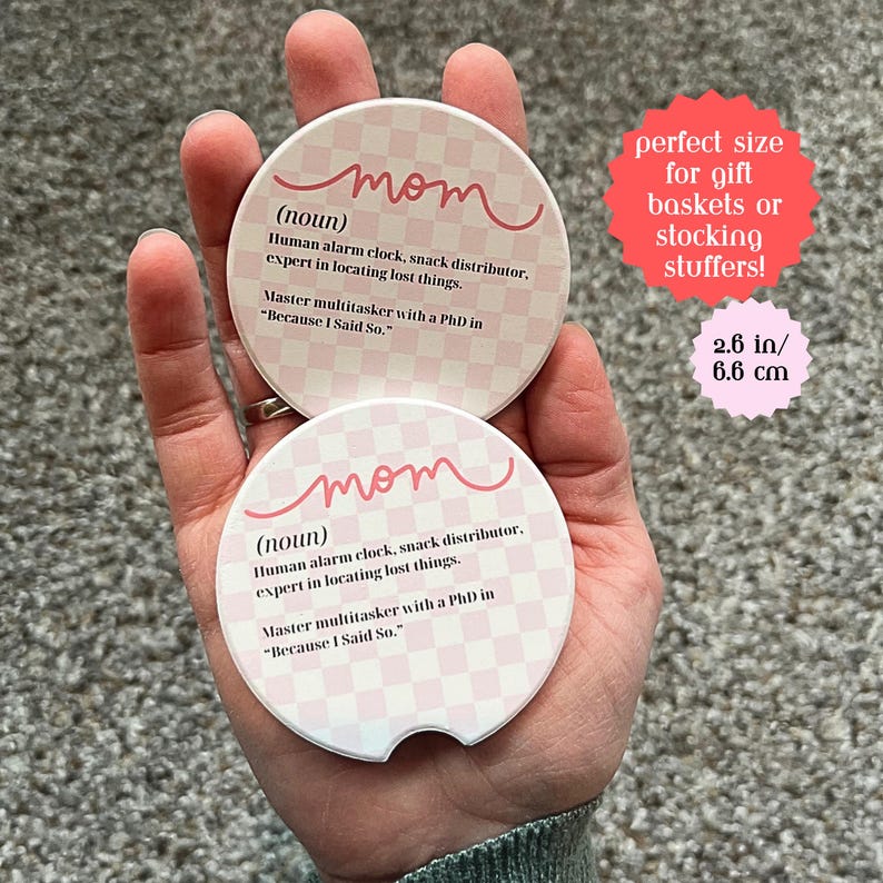 May include: Two round pink and white checkered coasters with the word "mom" written in pink cursive. The text below reads "(noun) Human alarm clock, snack distributor. expert in locating lost things. Master multitasker with a PhD in "Because I Said So."". The coasters are 2.6 inches in diameter.